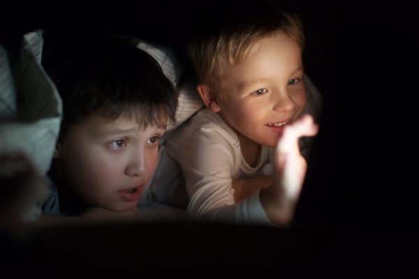 Two boys lying on bed under blanket at night. They watching movie or cartoon on pad. Screen enlighting their faces in darkness